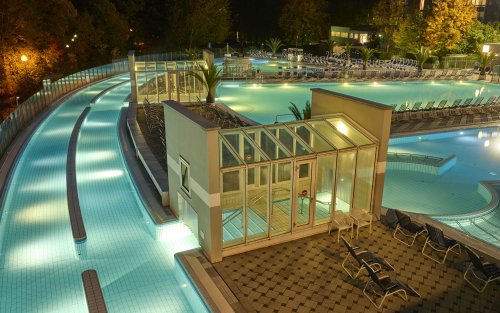 Nachtaufnahme eines beleuchteten Schwimmbads mit Rutschen und leeren Liegen.